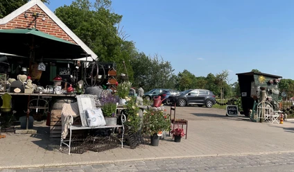 Hofladen Gartenshop Hof Saathoff Außenbereich eines Hofladens mit Pflanzen, Dekoartikeln, einer Bank und geparkten Autos im Hintergrund.