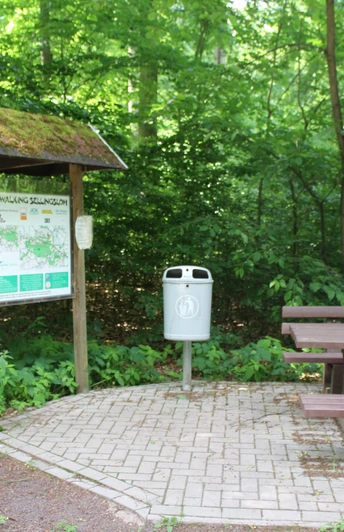 Eine beschilderte Infotafel für Nordic Walking neben einem Abfallbehälter und Holzbänken im Wald.
