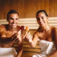Paar in der Sauna_Ishara_Foto_Michael Adamski.jpg Ein lächelndes Paar hält Eis am Stiel in einer Sauna, umwickelt mit weißen Handtüchern.