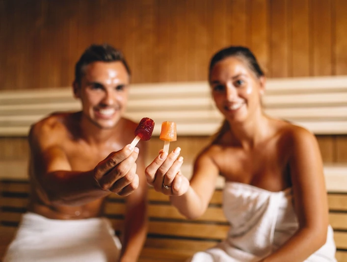 Paar in der Sauna_Ishara_Foto_Michael Adamski.jpg Ein lächelndes Paar hält Eis am Stiel in einer Sauna, umwickelt mit weißen Handtüchern.