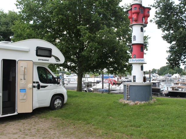 Wohnmobil neben rot-weißem Leuchtturm am Hafen, umgeben von Bäumen und Booten im Wasser.