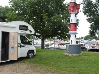 Wohnmobilstellplatz Marina Mehlbergen Wohnmobil neben rot-weißem Leuchtturm am Hafen, umgeben von Bäumen und Booten im Wasser.