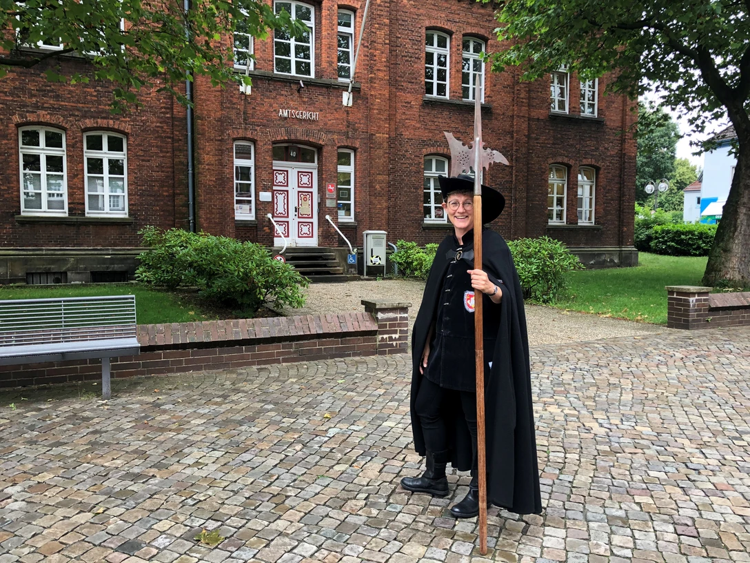 Nachtwächterin Martha van Acheim posiert mit Hellebarde vor einem historischen Backsteingebäude.
