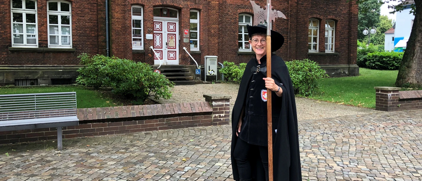 Nachtwächterin Martha van Acheim posiert mit Hellebarde vor einem historischen Backsteingebäude.