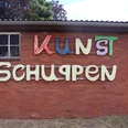 Bunte Buchstaben auf rotem Backstein, Holzfenster links, Kunstschuppen-Schriftzug mittig.