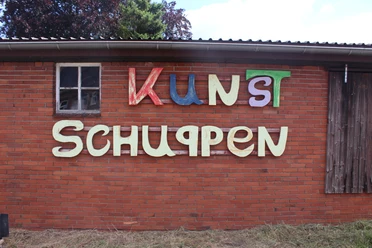 Kunstschuppen Asendorf Bunte Buchstaben auf rotem Backstein, Holzfenster links, Kunstschuppen-Schriftzug mittig.