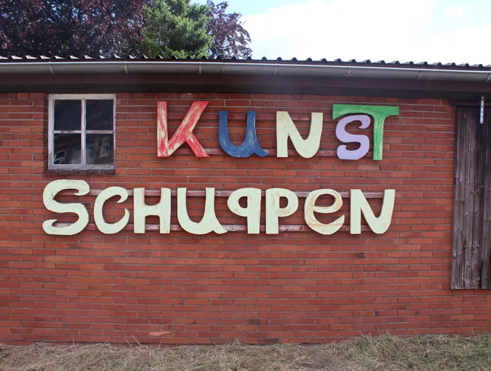 Kunstschuppen Asendorf Bunte Buchstaben auf rotem Backstein, Holzfenster links, Kunstschuppen-Schriftzug mittig.