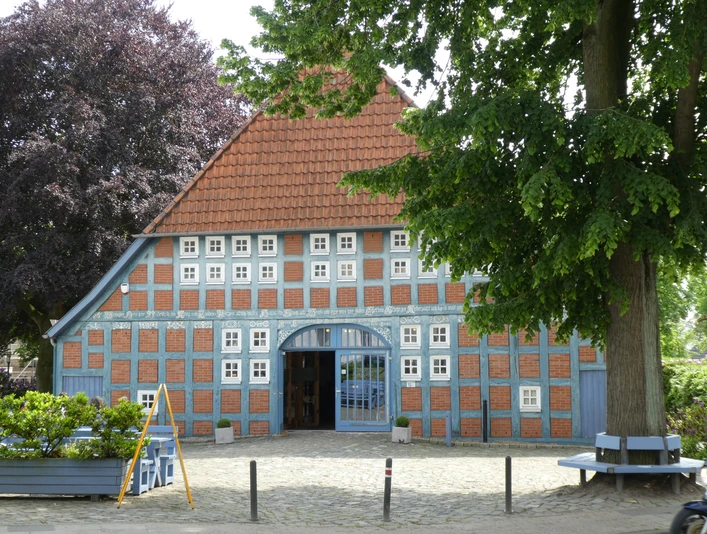 Historisches Fachwerkhaus mit blauen Fensterläden und rotem Ziegeldach unter grünen Bäumen.