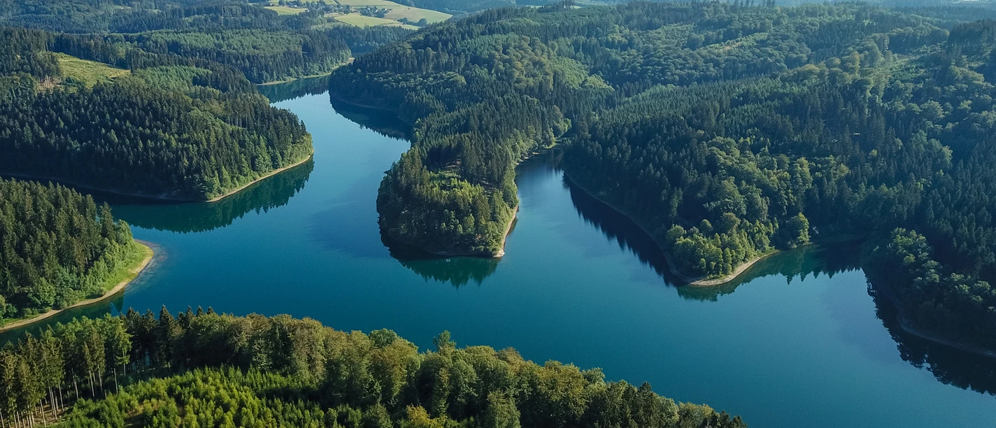 Genkeltalsperre <p>Luftaufnahme eines üppigen, grünen Waldes durchzogen von einem ruhigen, blauen Fluss, sanfte Hügel im Hintergrund.</p>