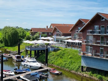 Hotel-Restaurant Bootshaus Hotel-Restaurant mit Terrassenschirmen am Fluss, umgeben von grüner Landschaft und Bootsstegen.