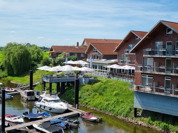 Hotel-Restaurant Bootshaus Hotel-Restaurant mit Terrassenschirmen am Fluss, umgeben von grüner Landschaft und Bootsstegen.