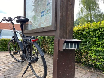 E-Bike-Ladestation Rathaus3.jpg Ein E-Bike lehnt an einer hölzernen Infotafel. Daneben ist eine Ladestation erkennbar.