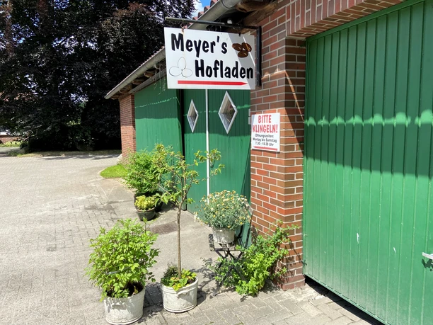 Meyers Hofladen.jpg Vor Meyers Hofladen grünen Pflanzen in Kübeln, flankiert von roten Backsteinwänden und grünen Toren.