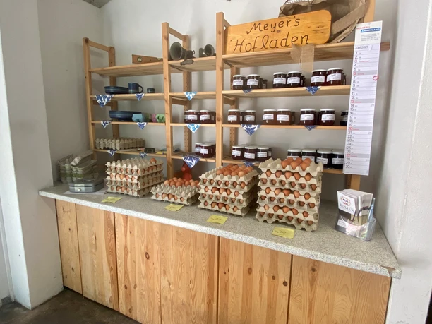 Meyers Hofladen2.jpg In einem gemütlichen Laden sind Holzbretterregale mit frischen Eiern und hausgemachten Marmeladen gefüllt.