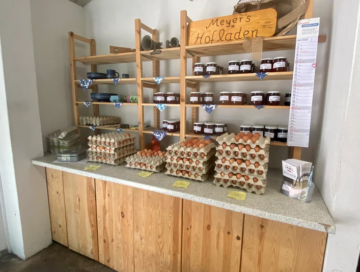Meyers Hofladen2.jpg In einem gemütlichen Laden sind Holzbretterregale mit frischen Eiern und hausgemachten Marmeladen gefüllt.