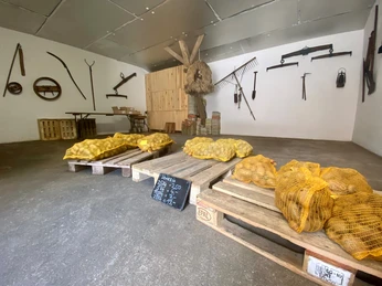 Meyers Hofladen3.jpg Ein rustikaler Hofladenraum mit landwirtschaftlichen Werkzeugen an der Wand. Auf Paletten liegen Kartoffelsäcke.