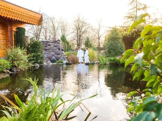 Teutoburger Wald-2015-Wellness-013-Teutoburger-Wald-Tourismus-D-Ketz.jpg Zwei Personen in weißen Bademänteln spazieren an einem Teich in einer grünen Gartenlandschaft.