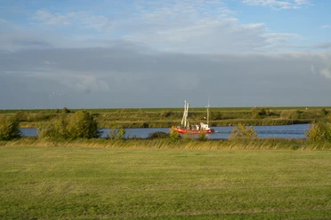 Krabbenkutter am Badesee Greetsiel Roter Krabbenkutter auf dem Wasser am Badesee Greetsiel, umgeben von Deich und Wiesen.