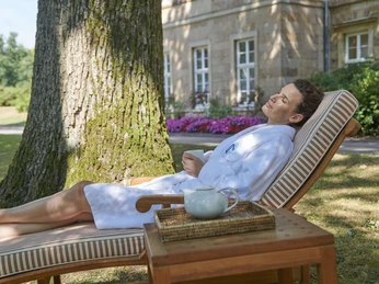 Gräflicher Park Health & Balance Resort Eine Person in einem weißen Bademantel entspannt auf einer Sonnenliege im schattigen Park vor einem Gebäude.