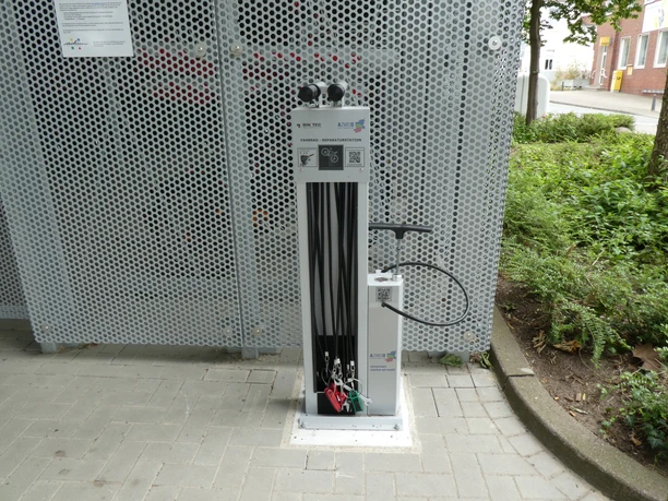 Lufttankstelle Bike & Ride Achimer Bahnhof Fahrrad-Luftstation am Achimer Bahnhof mit integrierter Pumpe, umgeben von Metallgitter und Pflanzen.