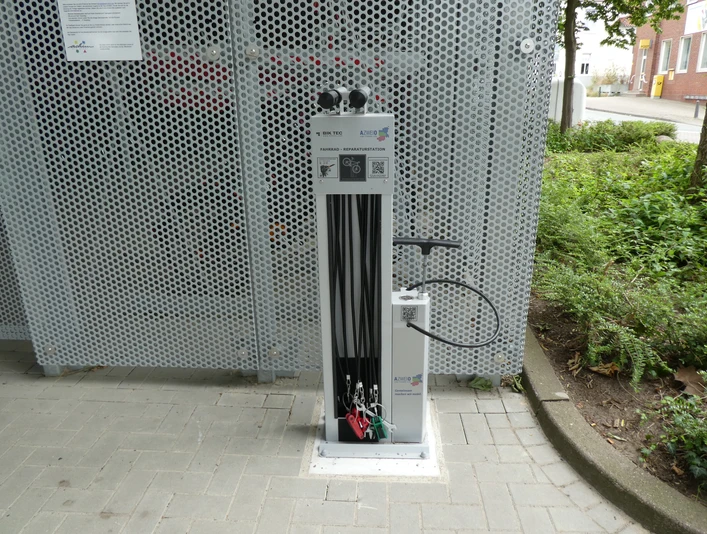Lufttankstelle Bike & Ride Achimer Bahnhof Fahrrad-Luftstation am Achimer Bahnhof mit integrierter Pumpe, umgeben von Metallgitter und Pflanzen.