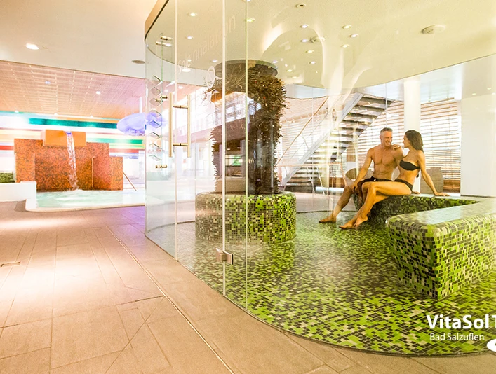 Soleinhalation VitaSol Therme Zwei Personen entspannen in der gläsernen Soleinhalation der VitaSol Therme auf grünen Mosaiksitzen.