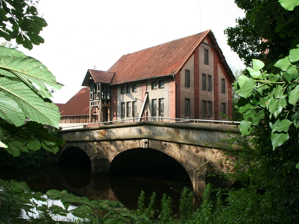 A historic bridge in front of a reddish-brown, multi-story building, nestled among lush, green foliage.