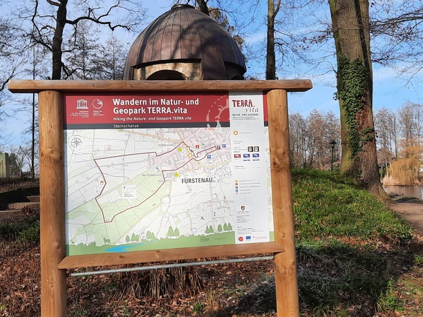 TafelFürstenauerPattSchlosspark.jpg Houten plank met wandelkaart in het natuurpark, omringd door bomen onder een heldere hemel.