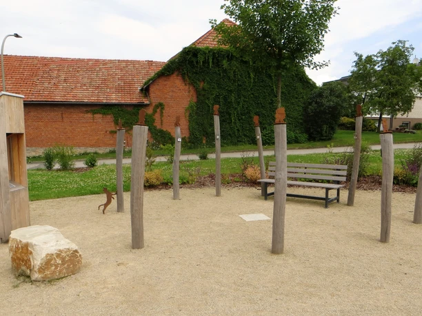 Ludowinengarten mit hölzernen Skulpturen und Sitzgelegenheit, umgeben von begrünten Gebäuden.