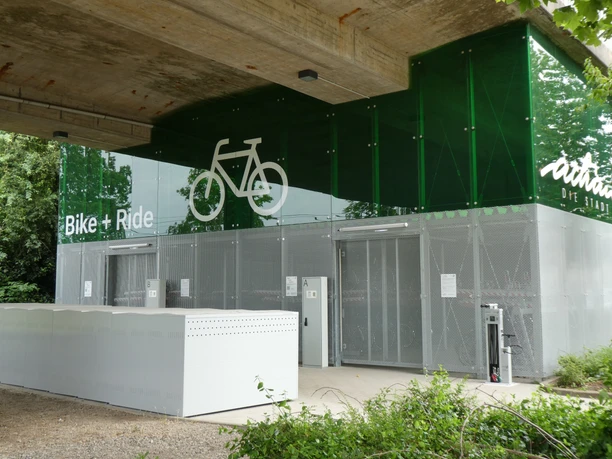 Moderne Fahrradanlage unter Brückenkonstruktion mit grünem Glasdach und Fahrradpiktogramm.