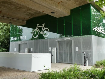 Bike & Ride Station Achim Moderne Fahrradanlage unter Brückenkonstruktion mit grünem Glasdach und Fahrradpiktogramm.
