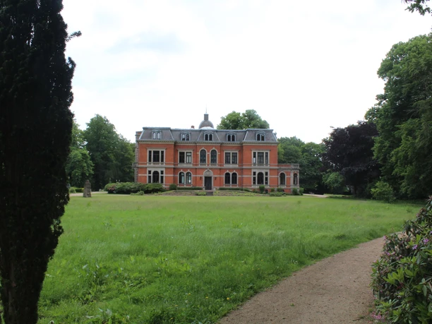Red brick building in the middle of a spacious park with a well-kept lawn and surrounding trees.