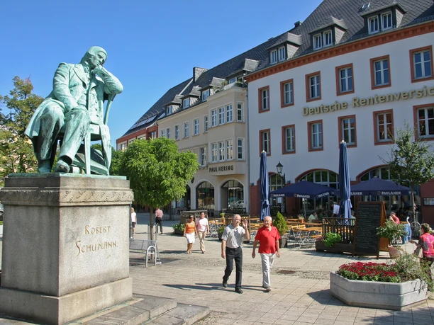007 - Robert Schumann Denkmal am Hauptmarkt - © Kultour Z.jpg Bronzestatue von Robert Schumann in nachdenklicher Pose auf einem Platz vor historischen Gebäuden.