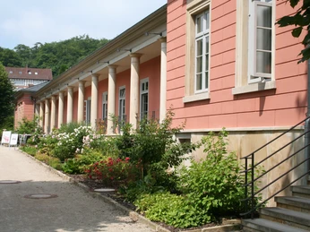 Romantik Bad Rehburg Historisches Gebäude mit roten Wänden und Säulen, flankiert von einem gepflegten Blumenbeet.Historic building with red walls and columns, flanked by a well-tended flower bed.Historisk bygning med røde mure og søjler, flankeret af et velplejet blomsterbed.Historisch gebouw met rode muren en zuilen, geflankeerd door een goed onderhouden bloembed.