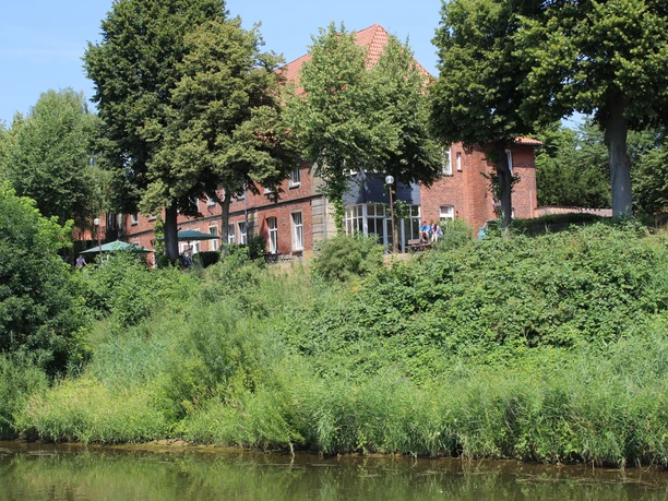A 19th-century red-brick building surrounded by lush greenery on the banks of a river.