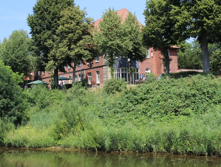 Ein rotes Backsteingebäude aus dem 19. Jahrhundert umgeben von üppigem Grün am Ufer eines Flusses.