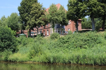 Hasbergscher Hof Ein rotes Backsteingebäude aus dem 19. Jahrhundert umgeben von üppigem Grün am Ufer eines Flusses.
