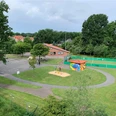 Außenansicht CASA Das Bild zeigt einen kleinen Spielplatz mit Sandkasten und bunter Hütte zwischen Bäumen, nahe einem roten Backsteingebäude und einem eingezäunten Sportplatz.