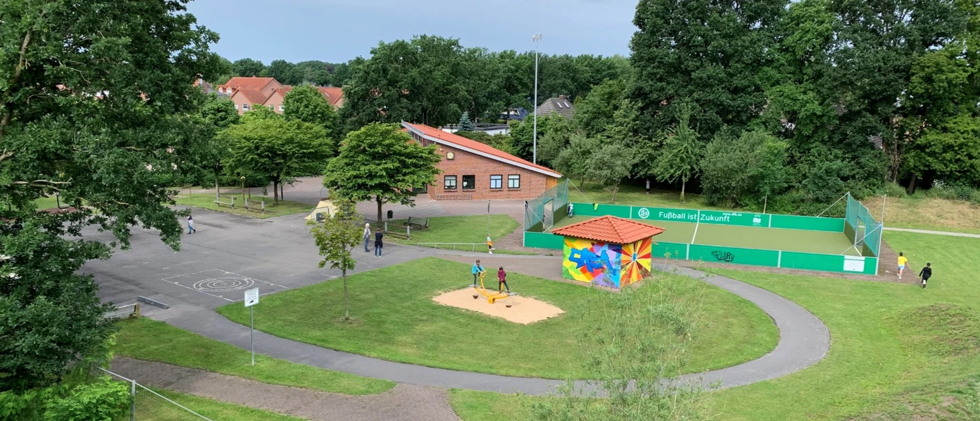 Außenansicht CASA Das Bild zeigt einen kleinen Spielplatz mit Sandkasten und bunter Hütte zwischen Bäumen, nahe einem roten Backsteingebäude und einem eingezäunten Sportplatz.