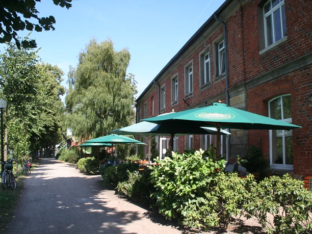 A brick building with green sunshades; café patrons are sitting relaxed on a path in the outdoor seating area.