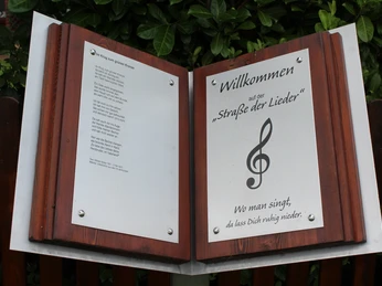 Ein Willkommensschild der "Straße der Lieder" mit Notensymbol steht vor einer grünen Hecke.