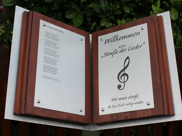 Ein Willkommensschild der "Straße der Lieder" mit Notensymbol steht vor einer grünen Hecke.