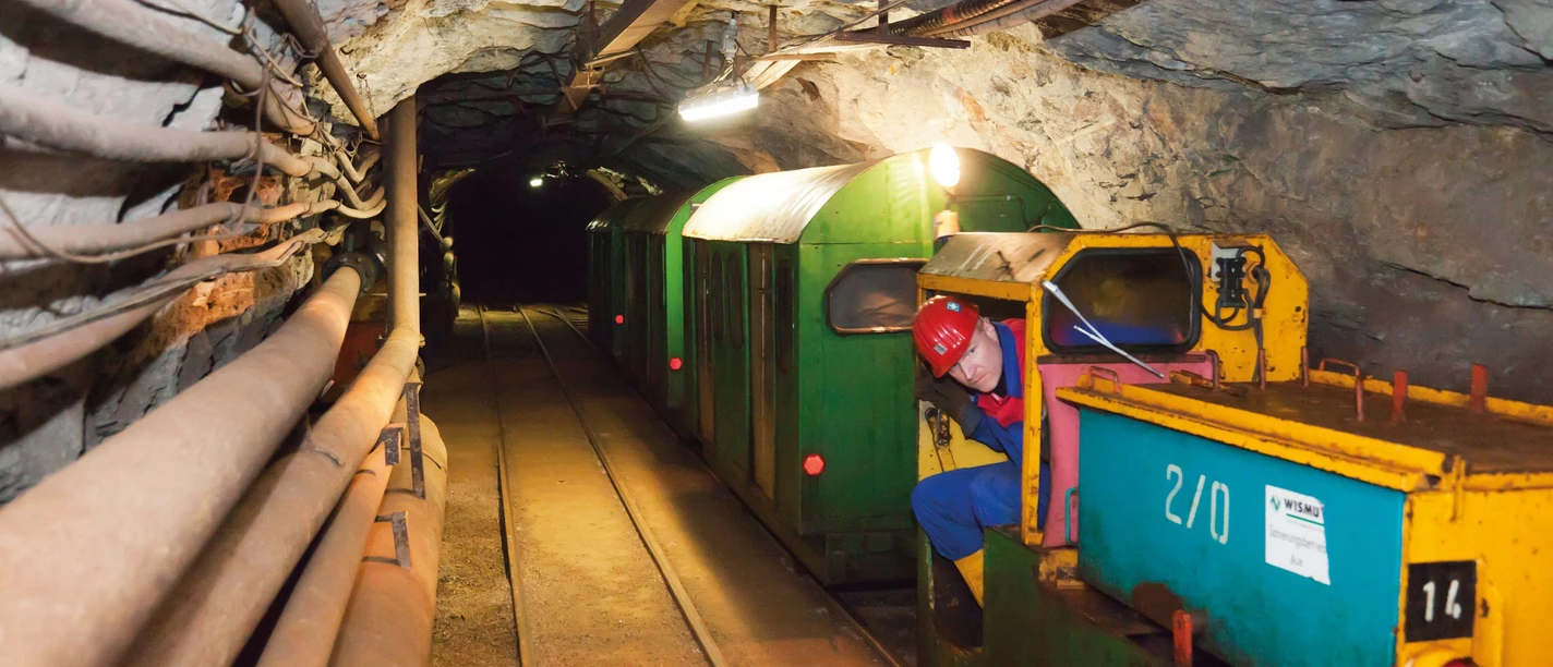 Visitor train runs through illuminated mine tunnel, worker in blue outfit controls locomotive.