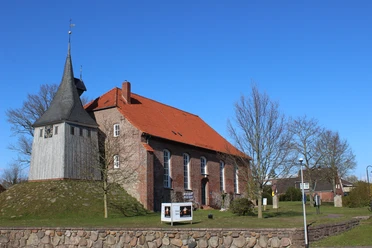 Backsteinkirche mit rotem Dach und freistehendem Holzturm auf einem Hügel unter blauem Himmel.