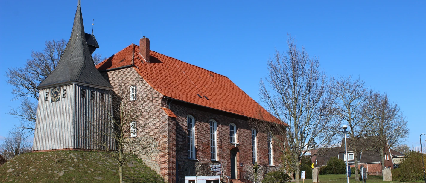 Backsteinkirche mit rotem Dach und freistehendem Holzturm auf einem Hügel unter blauem Himmel.