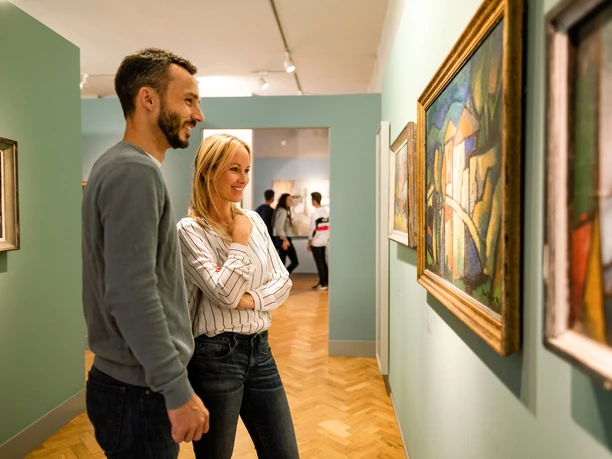 A smiling couple looks at paintings in an art gallery with parquet flooring.