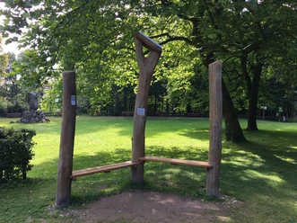 Hörstation am Hermannsdenkmal Holzbank in einer grünen Parklandschaft, umgeben von Bäumen, lädt zur Rast und zum Lauschen ein.