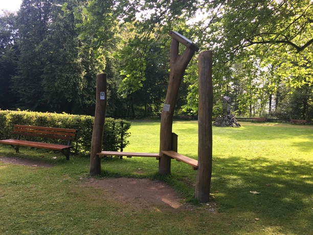 Hörstation aus Holz im Grünen mit Umgebung aus Bäumen und einer Parkbank im Hintergrund.