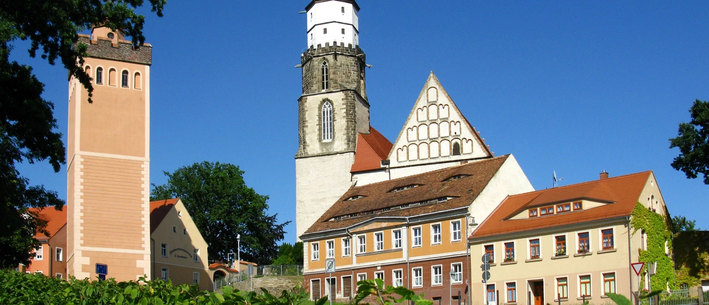 Red tower and main church with half-timbered façade in green surroundings, under a blue sky.