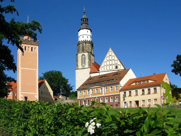 Red tower and main church with half-timbered façade in green surroundings, under a blue sky.